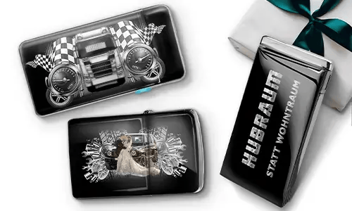 Drei schwarze Geldbörsen mit Auto-Designs und karierten Flaggen, vor einem weißen Hintergrund. Eine zeigt ein Auto mit Rädern, eine andere ein Paar, und eine trägt den Text „Hubraum statt Wohntraum“.